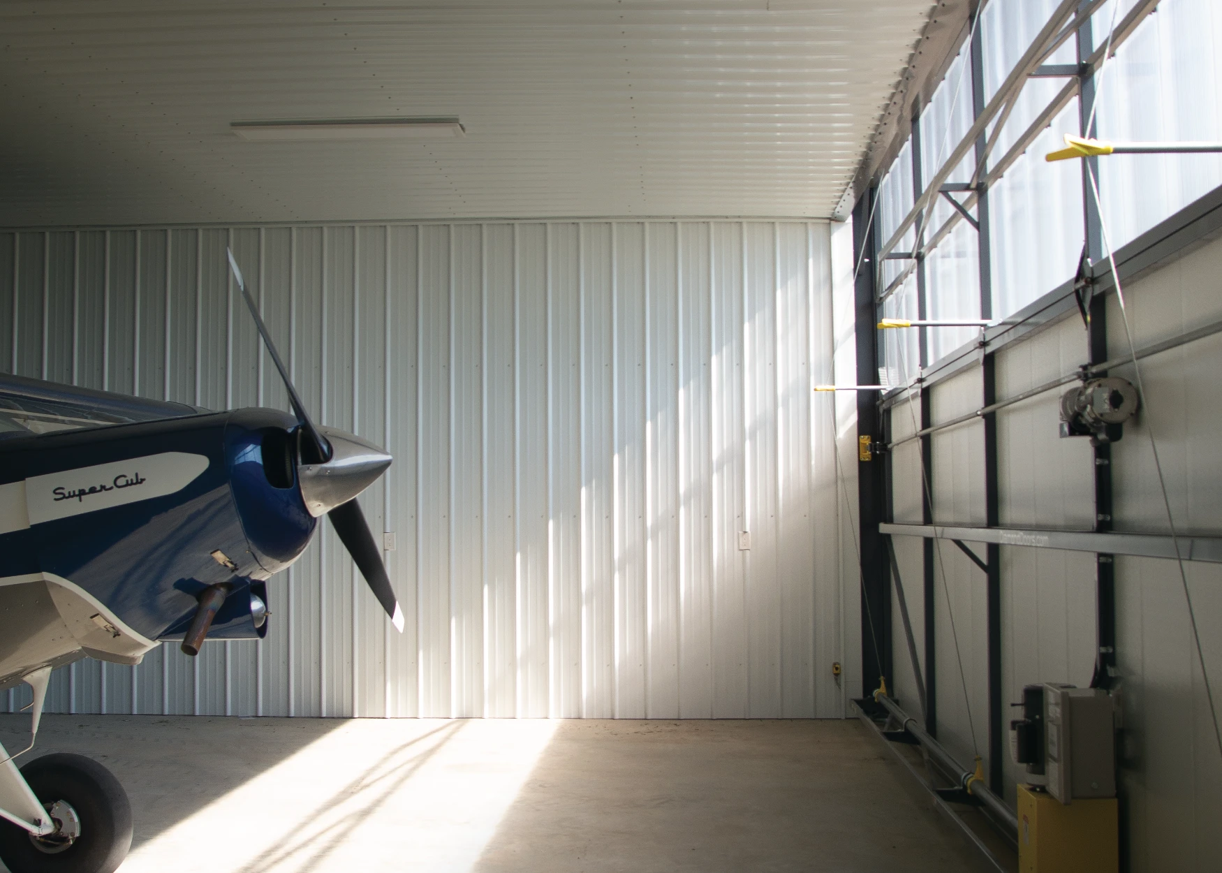 A Supercub airplane inside a hangar with a bifold door and polycarbonate sheeting.